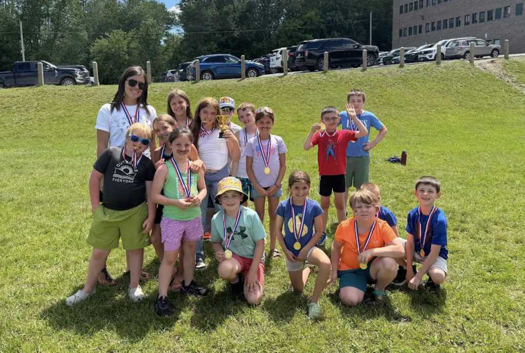 A teacher stands with a group of medal-winning students on a grassy hillside near a school parking area. One student holds a trophy while others wear medals and smile for the camera.