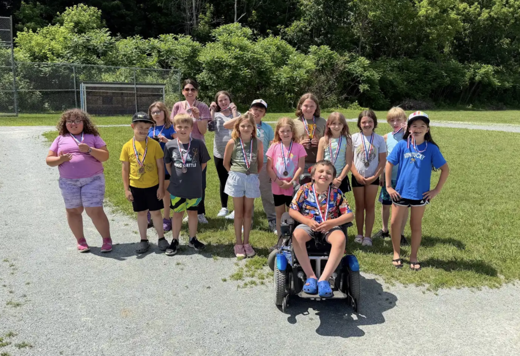 A group of students wearing medals poses with a teacher outdoors on a field. One student sits in a motorized wheelchair at the front while classmates smile around them, celebrating their accomplishments.