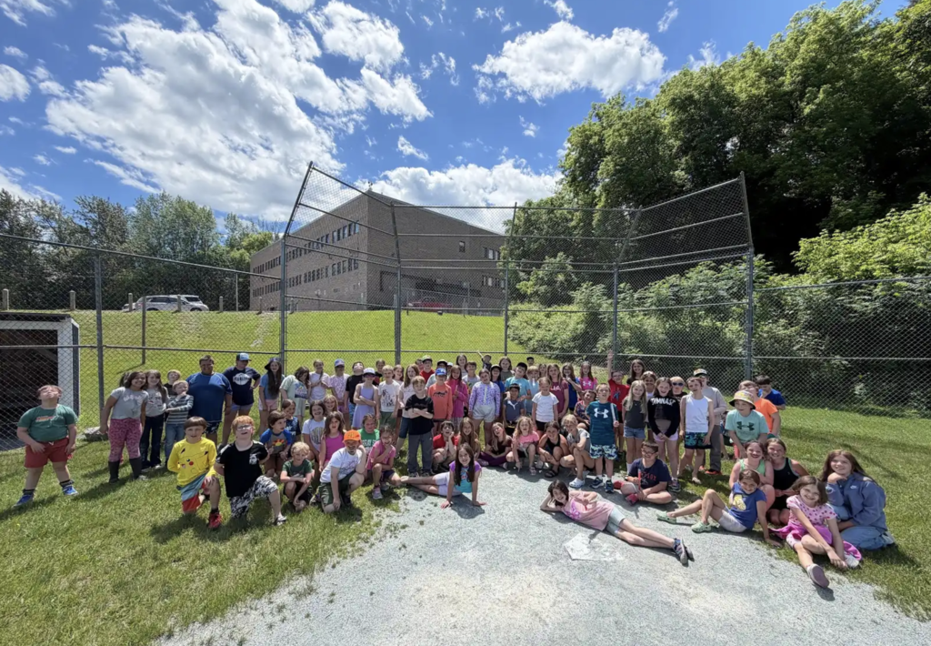 A large group of elementary students gathers on a baseball field under a bright blue sky with scattered clouds. Students pose together in front of the backstop fence during an outdoor field day celebration.