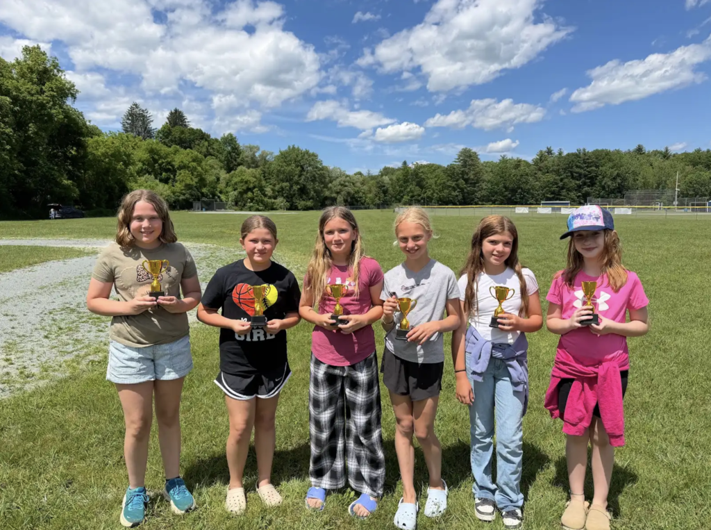 Six elementary students stand in a row on a grassy field holding small gold trophies. Trees and athletic fields are visible in the background on a sunny day.