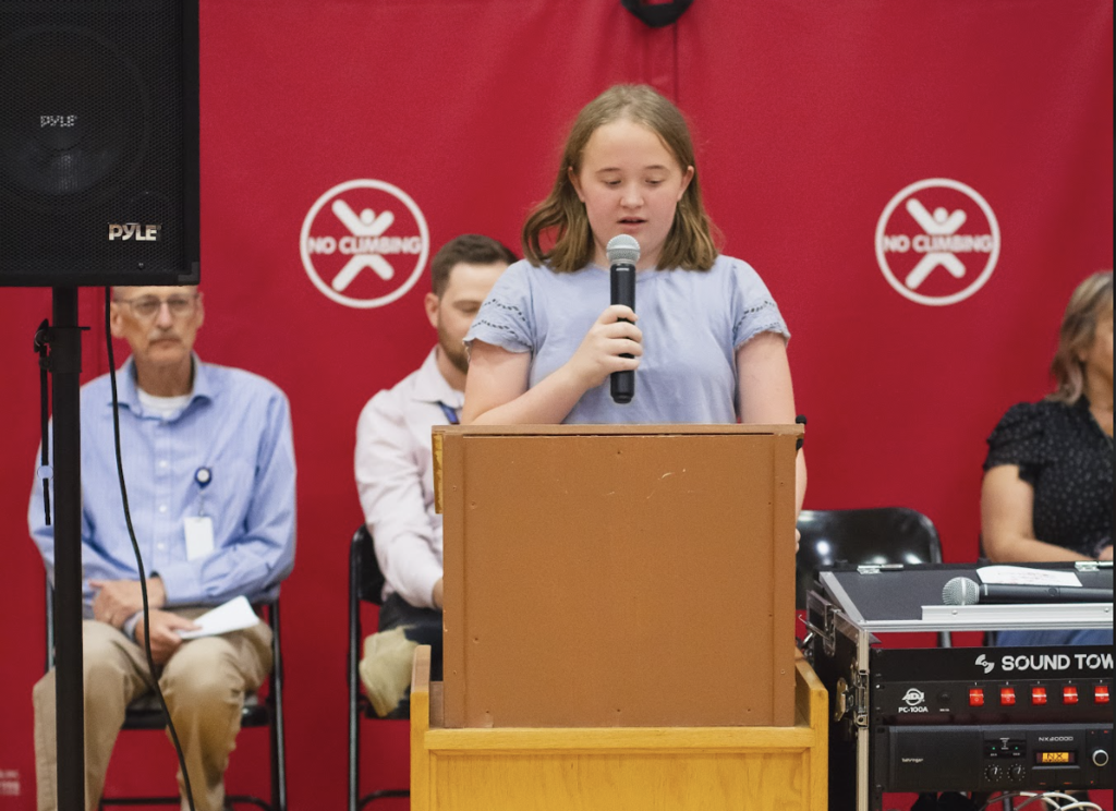 A sixth-grade student stands at a podium speaking into a microphone during the Castleton Elementary School Grade 6 Torch Ceremony while school administrators and staff sit behind her.
