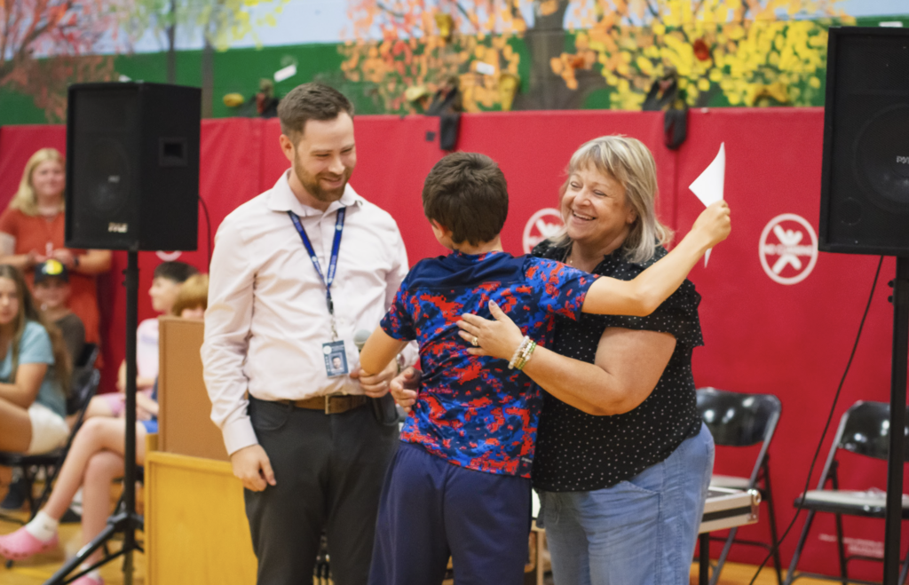 A young student hugs their teacher as Principal Jared Danyow smiles nearby during the torch-passing portion of the ceremony.