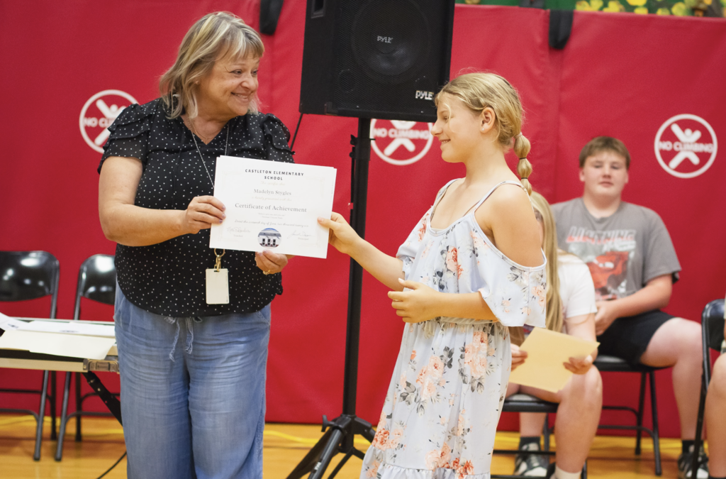 A teacher presents a Certificate of Achievement to a smiling sixth-grade student during the Grade 6 Torch Ceremony.