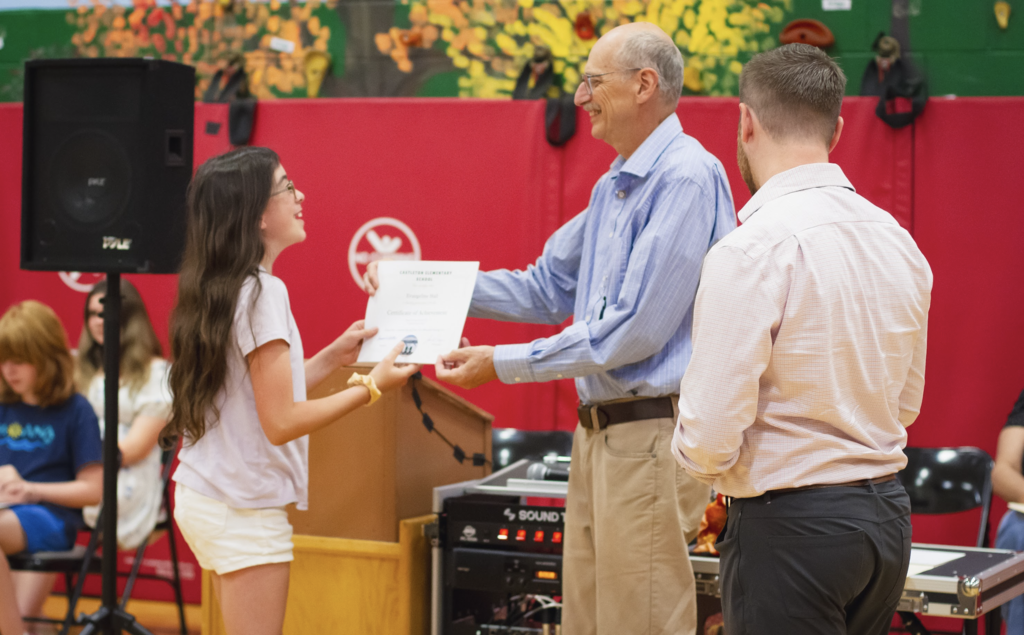 A sixth-grade student receives a certificate from their teacher while Principal Danyow looks on during the recognition ceremony.