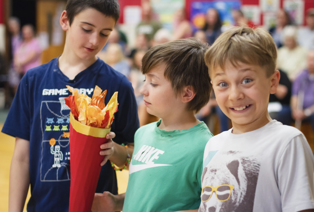 Three students hold the ceremonial torch together while one student smiles playfully toward the camera during the Grade 6 Torch Ceremony.