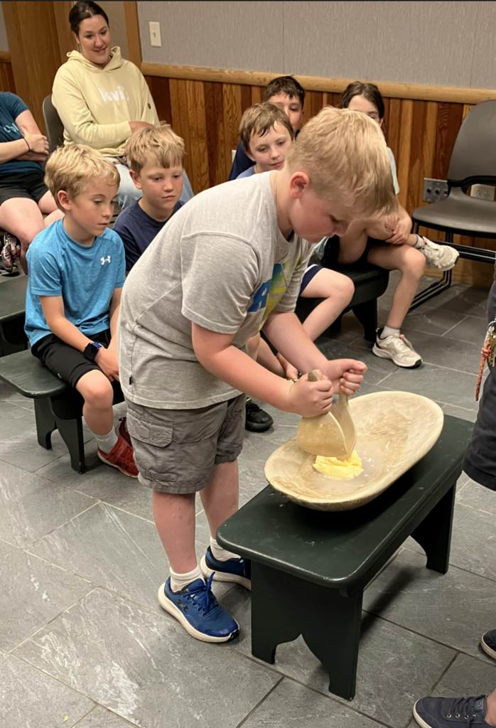 A second grade student uses a traditional wooden butter paddle to shape freshly made butter while classmates watch from nearby benches.