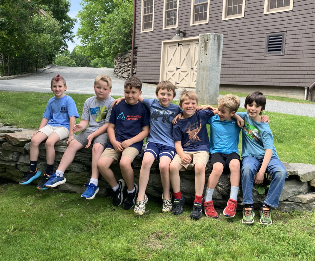 Seven second grade boys sit together on a stone wall outside a farm building, smiling and posing for a group photo during their field trip.