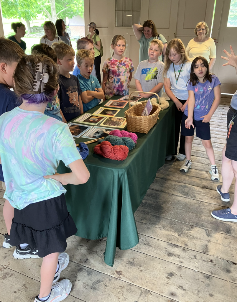 Students gather around a display table covered with wool, yarn, and educational materials as a Billings Farm educator explains how wool is processed and used.