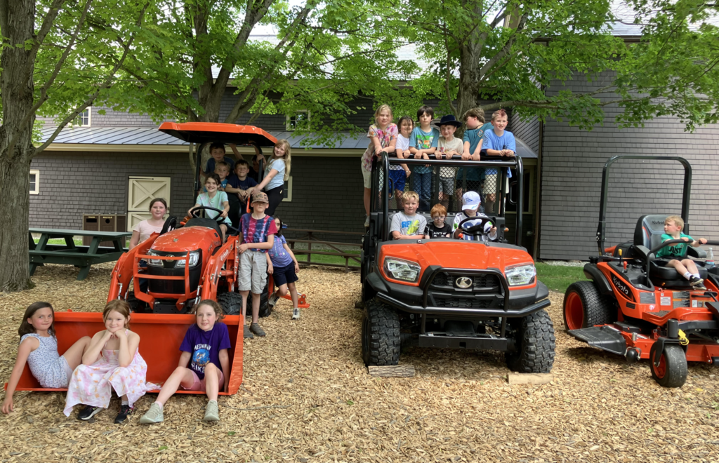 A large group of second graders poses on and around bright orange farm equipment, including tractors and utility vehicles, beneath shady trees at Billings Farm.