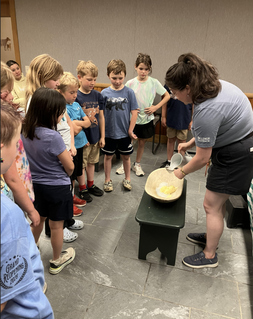 A Billings Farm educator demonstrates the butter-making process in a large wooden bowl while a group of Castleton Elementary second graders gathers closely around to watch and learn.
