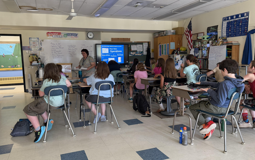 Students sit at desks in a classroom during Move Up Day at Fair Haven Grade School. A teacher stands at the front of the room leading a lesson while students listen and look toward an interactive display screen. The classroom is decorated with educational posters, bookshelves, and student materials as fourth graders get a preview of fifth grade.