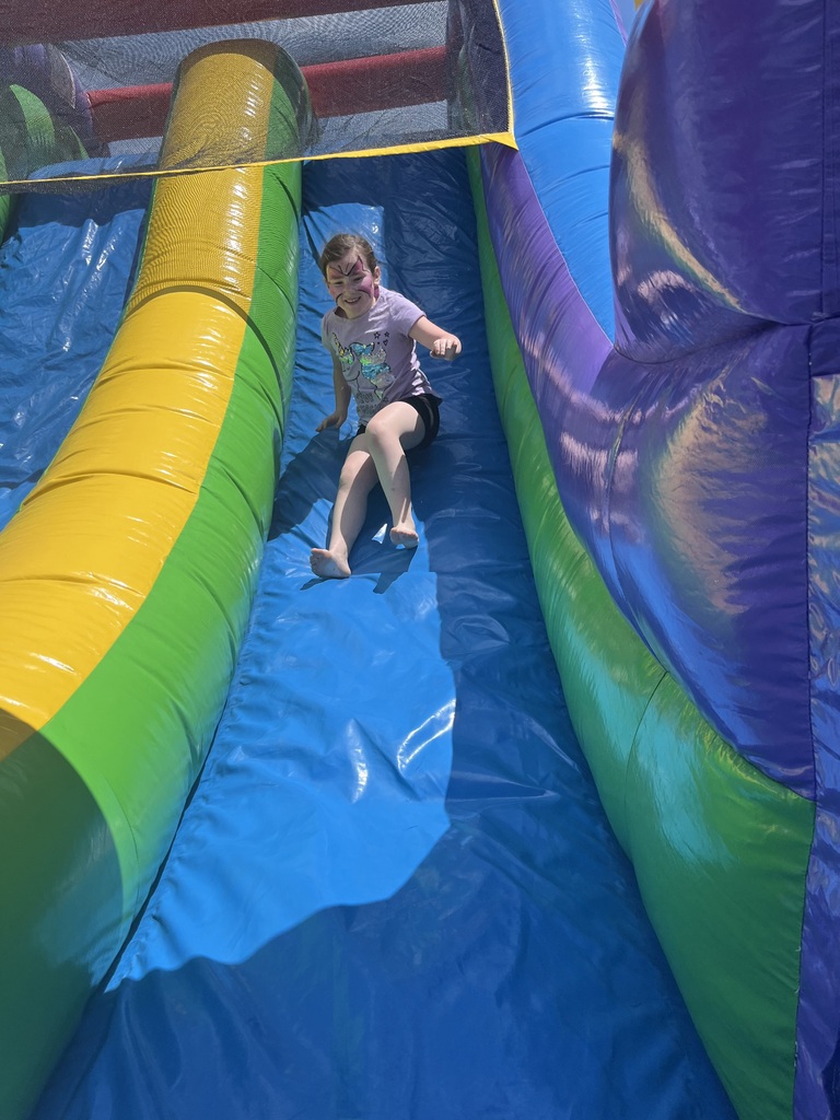 A young girl with her face painted slides down a large, colorful blue and green inflatable water slide on a bright, sunny day. I