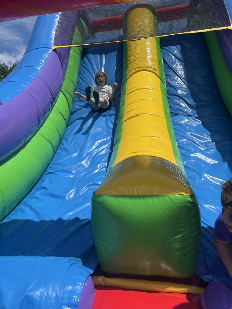A young child with an excited, wide-open expression slides feet-first down a tall, vibrant inflatable slide under a clear blue sky. Sack Race Activity (Wide Shots) IMG