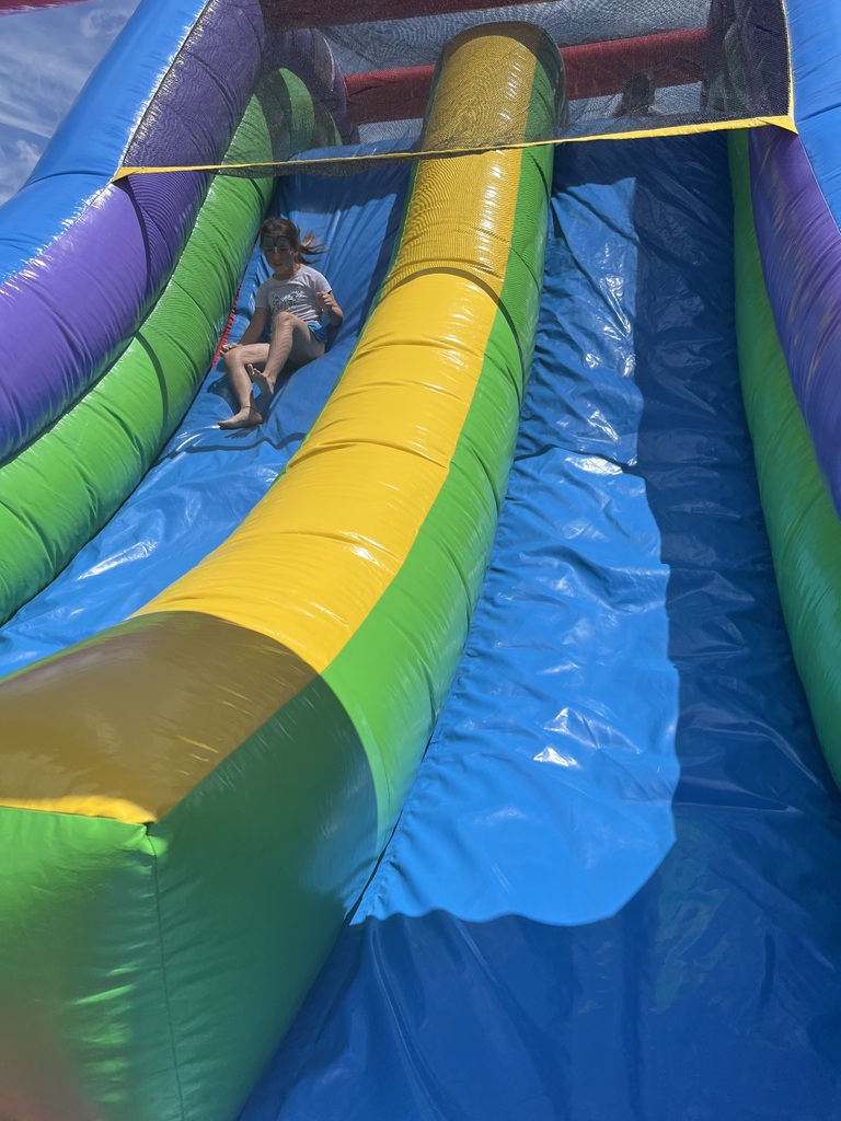 A young girl with her face painted slides down a large, colorful blue and green inflatable water slide on a bright, sunny day.