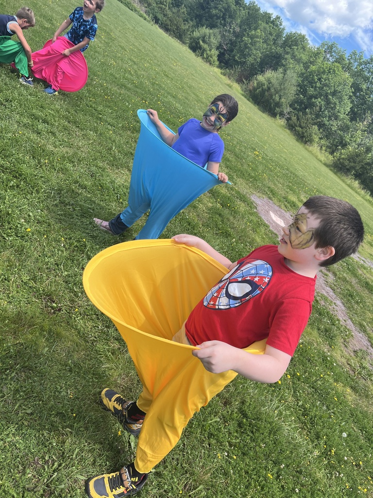 Children participate in a field day game on a large grassy lawn, some wearing colorful giant sacks while others throw and catch a beach ball. IMG_6229.jpg: A wide shot of a field day event where children in brightly colored racing sacks compete on a vast, green grass field under a blue sky with fluffy white clouds.