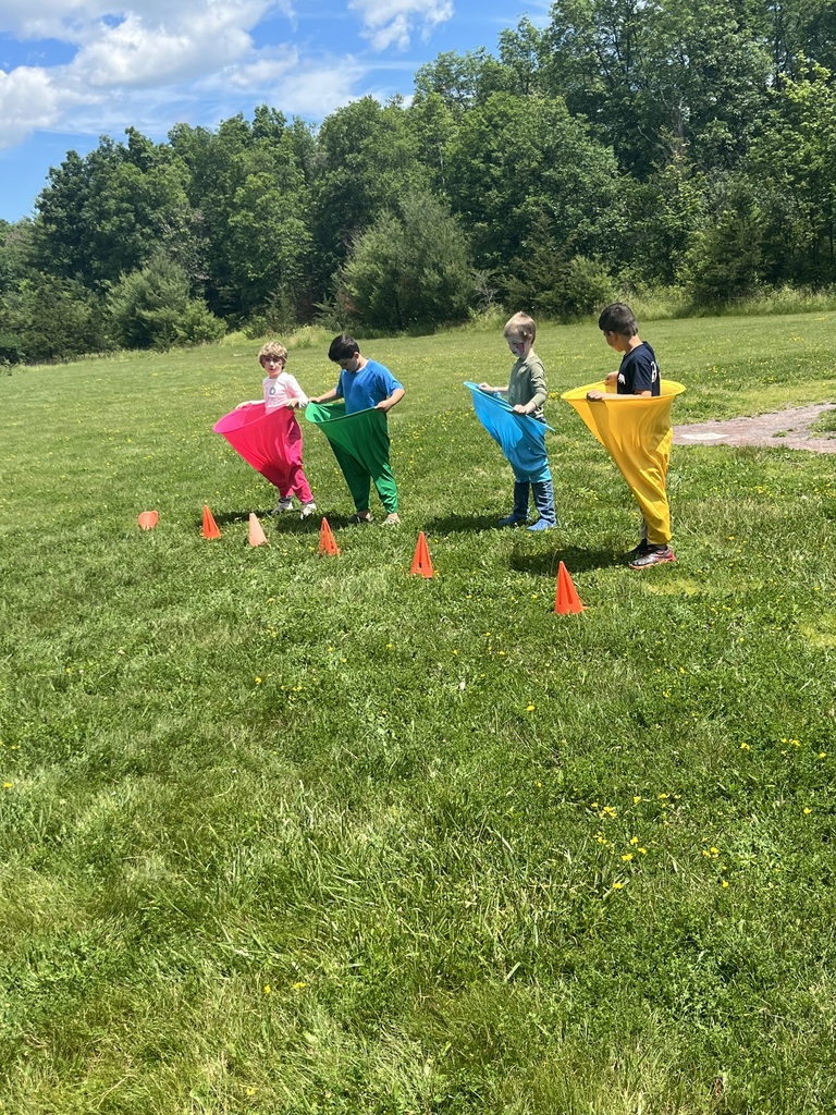 Four young children stand in a grassy field lined up behind orange cones, each stepped inside a brightly colored, oversized fabric sack (pink, green, blue, and yellow) preparing for a race.