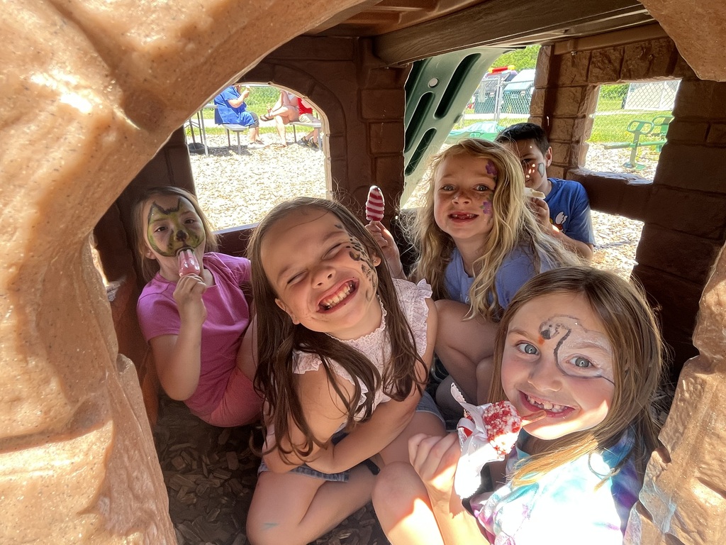 A group of five smiling children with painted faces squeeze into a small plastic playhouse on a playground while enjoying colorful popsicles and ice cream. Picnic Tables & Outdoor Treats IMG_6243.jpg: A group of children with face paint sit around a grey picnic table under a blue umbrella, eating cold treats on a sunny day near a yellow playground slide. IMG_6250.jpg: A smiling girl wearing a black "Boys & Girls Club of Rutland County" t-shirt proudly holds up a red, white, and blue striped popsicle while standing on a wood-chipped playground.