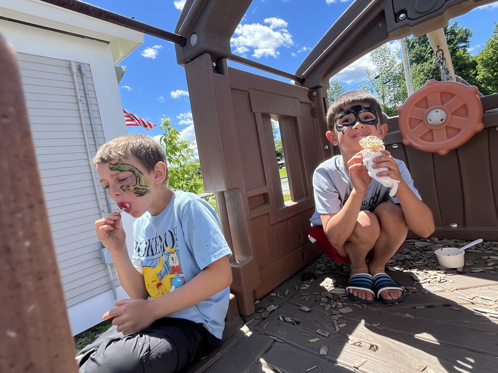 : Two young boys with superhero face paint take a break inside a wooden playground playhouse, one eating a popsicle and the other enjoying an ice cream cone.