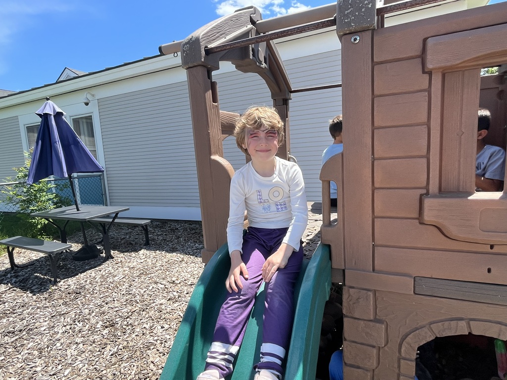 A child with subtle red face paint sits smiling at the bottom of a small green slide attached to a brown wooden playground structure. IMG_6248.jpg: Two young boys with superhero face paint take a break inside a wooden playground playhouse, one eating a popsicle and the other enjoying an ice cream cone.