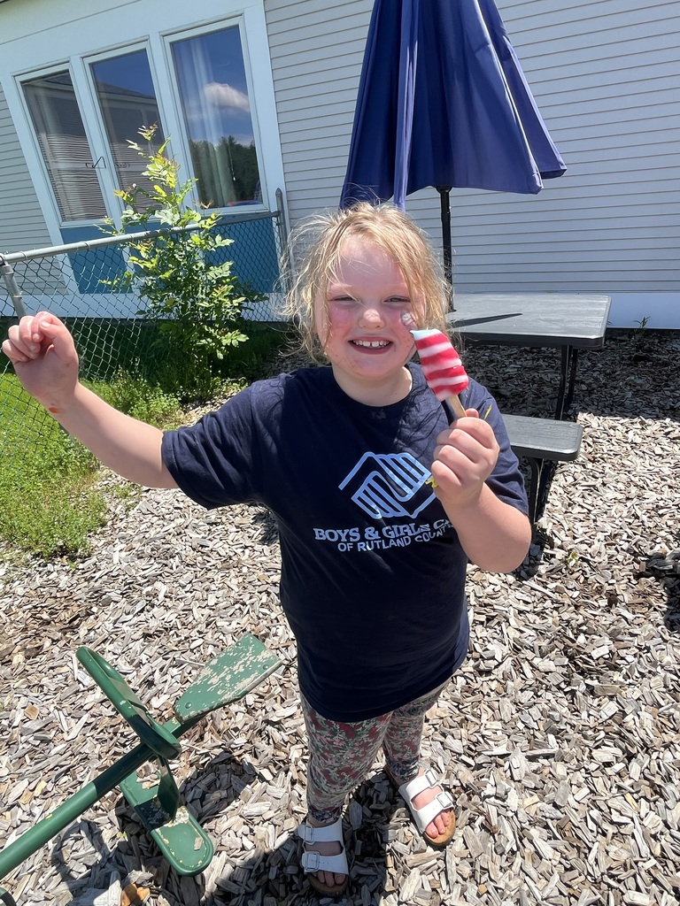 A smiling girl wearing a black "Boys & Girls Club of Rutland County" t-shirt proudly holds up a red, white, and blue striped popsicle while standing on a wood-chipped playground.