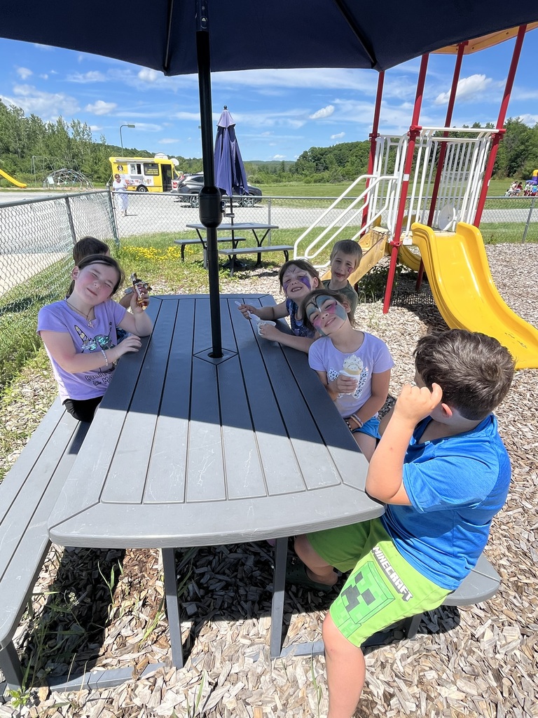 A group of five smiling children with painted faces squeeze into a small plastic playhouse on a playground while enjoying colorful popsicles and ice cream. Picnic Tables & Outdoor Treats IMG_6243.jpg: A group of children with face paint sit around a grey picnic table under a blue umbrella, eating cold treats on a sunny day near a yellow playground slide. IMG_6250.jpg: A smiling girl wearing a black "Boys & Girls Club of Rutland County" t-shirt proudly holds up a red, white, and blue striped popsicle while standing on a wood-chipped playground.