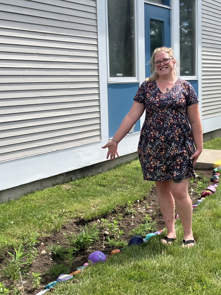 A smiling woman with glasses and blonde hair wearing a floral dress stands outside next to a newly planted garden bed. She gestures proudly with an open hand toward a long line of colorful, hand-painted rocks that border the edge of the soil. The garden runs along the side of a grey siding building with blue trim around a door and windows.