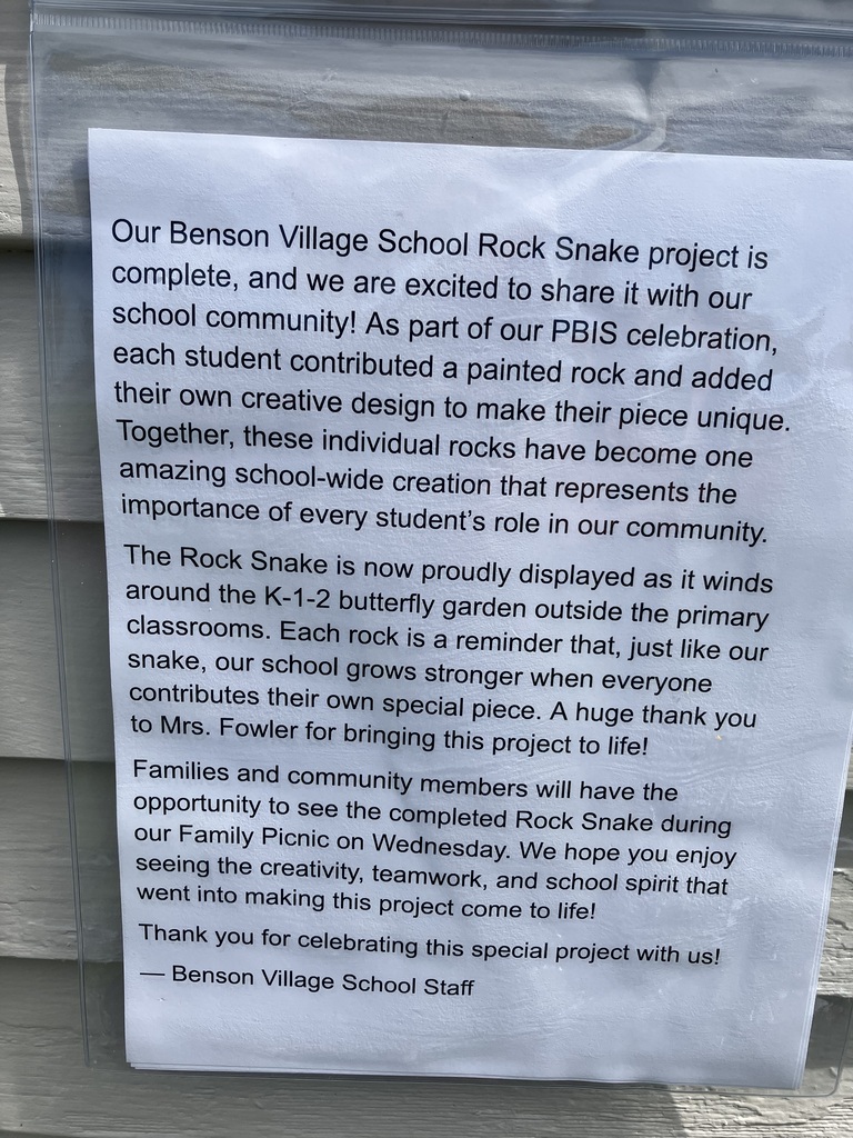 A close-up of a printed announcement from the Benson Village School Staff, protected inside a clear plastic sleeve and posted on a grey siding wall. The text announces the completion of the "Benson Village School Rock Snake project" as part of a PBIS celebration, noting that every student contributed a unique painted rock. It explains that the snake winds around the K-1-2 butterfly garden, thanks Mrs. Fowler for bringing the project to life, and invites families to view it during the Wednesday Family Picnic.