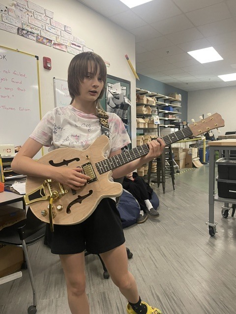 A student stands in a classroom holding and playing a handmade electric guitar. The unfinished natural wood guitar features gold hardware and a custom design. Shelving, tools, and classroom materials are visible in the background, highlighting the STEM and design setting where the student completed the project.