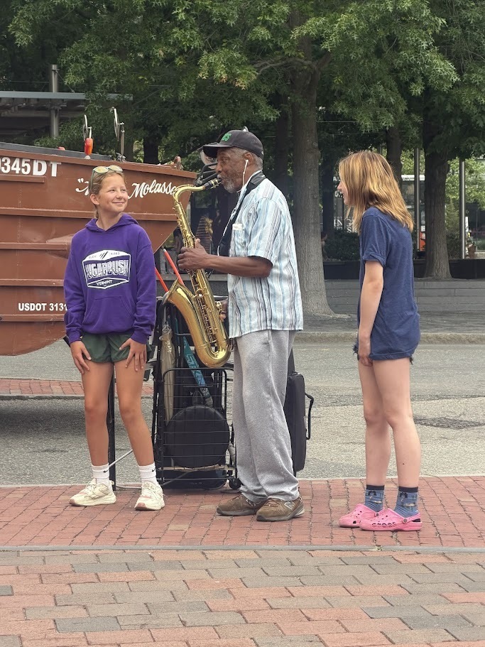 Two students stand smiling on a city sidewalk while listening to a street musician play a saxophone. Trees, brick pavement, and city buildings are visible in the background.