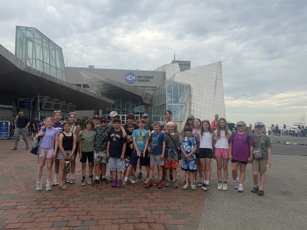A group of students and chaperones pose for a photo outside the New England Aquarium in Boston. The aquarium's distinctive glass architecture and waterfront setting are visible behind them.