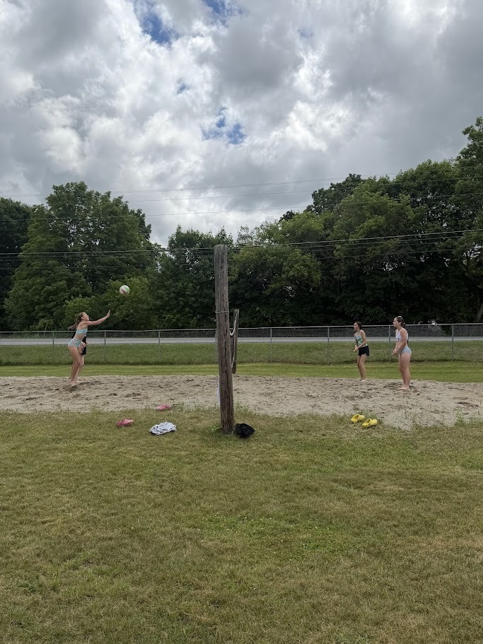 Students play a game of sand volleyball on an outdoor court near a lake. A volleyball is in the air above the net while players prepare for the next hit under a cloudy summer sky.