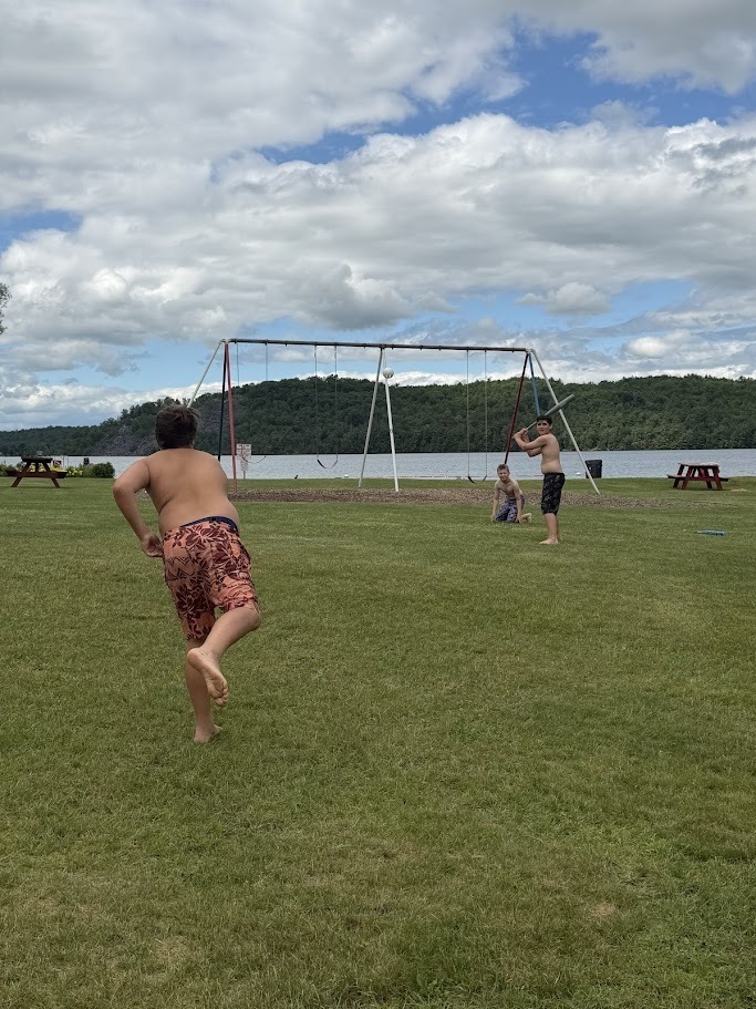 Students play a game of baseball on a grassy field beside a lake. One student runs toward home plate while another prepares to field the ball near a swing set in the background.