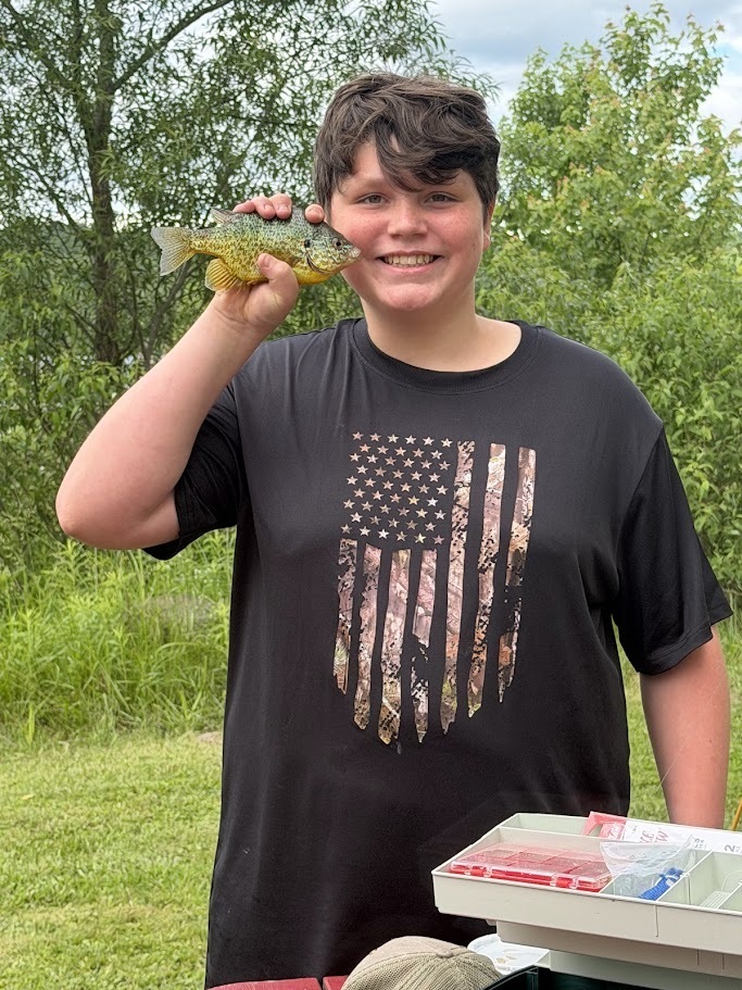 A smiling student proudly holds up a freshly caught sunfish while standing outdoors near a lakeside fishing area. A tackle box and fishing gear are visible nearby.