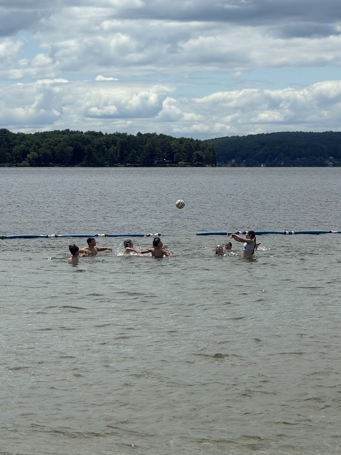 Students play a game with a beach ball while standing in waist-deep water at a lakeside swimming area. The ball is suspended in the air above the group.