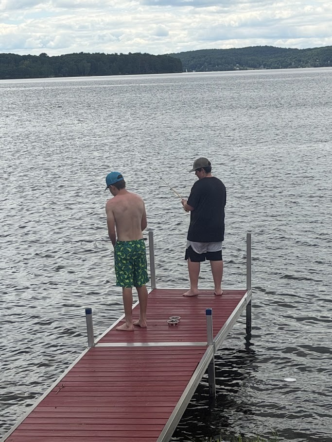 Two students fish from the end of a red dock extending into a lake. The students stand quietly with fishing rods while surrounded by calm water and distant wooded hills.