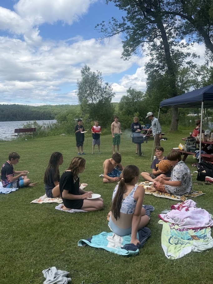 Students gather on blankets and lawn chairs for a picnic lunch beside a lake. Some students eat while others stand near a grill and canopy tent during an outdoor recreation day.