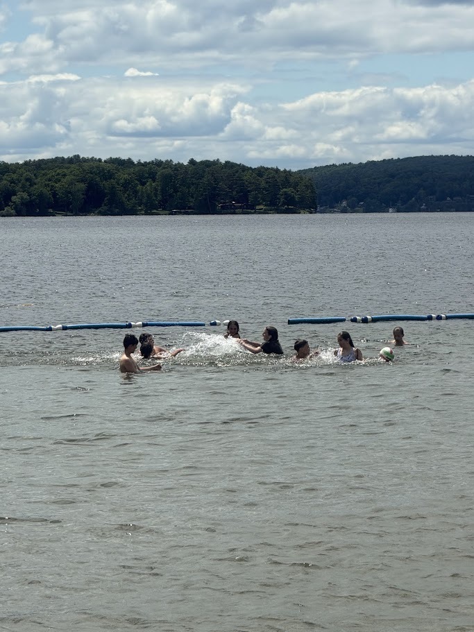 Students splash and play together in a designated swimming area of a lake. Floating lane markers and wooded shoreline are visible in the background.