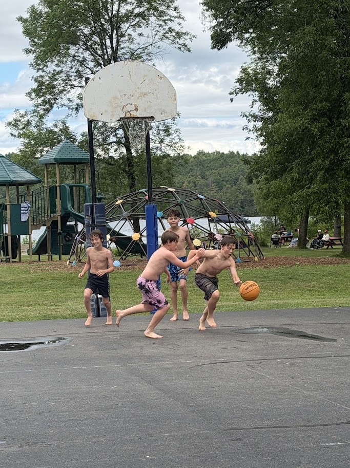 Four students play a barefoot game of basketball on an outdoor court near a playground and lakeside recreation area. One student dribbles the ball while others defend and watch the play.