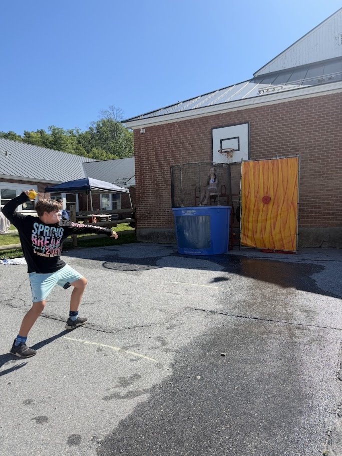 A student prepares to throw a ball at a dunk tank target during a school field day. A staff member sits above the tank while classmates watch nearby.
