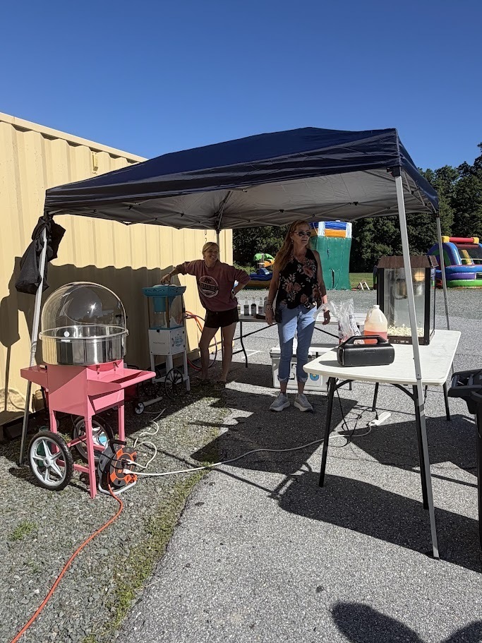 A cotton candy machine sits under a canopy tent during a school field day. Two adults stand nearby helping run the station while inflatable attractions can be seen in the background.
