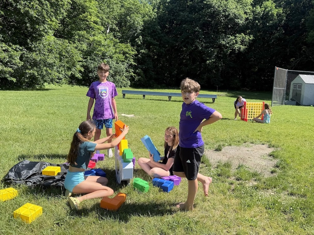 Students work together to build structures with oversized colorful foam blocks on the grass during outdoor field day activities.