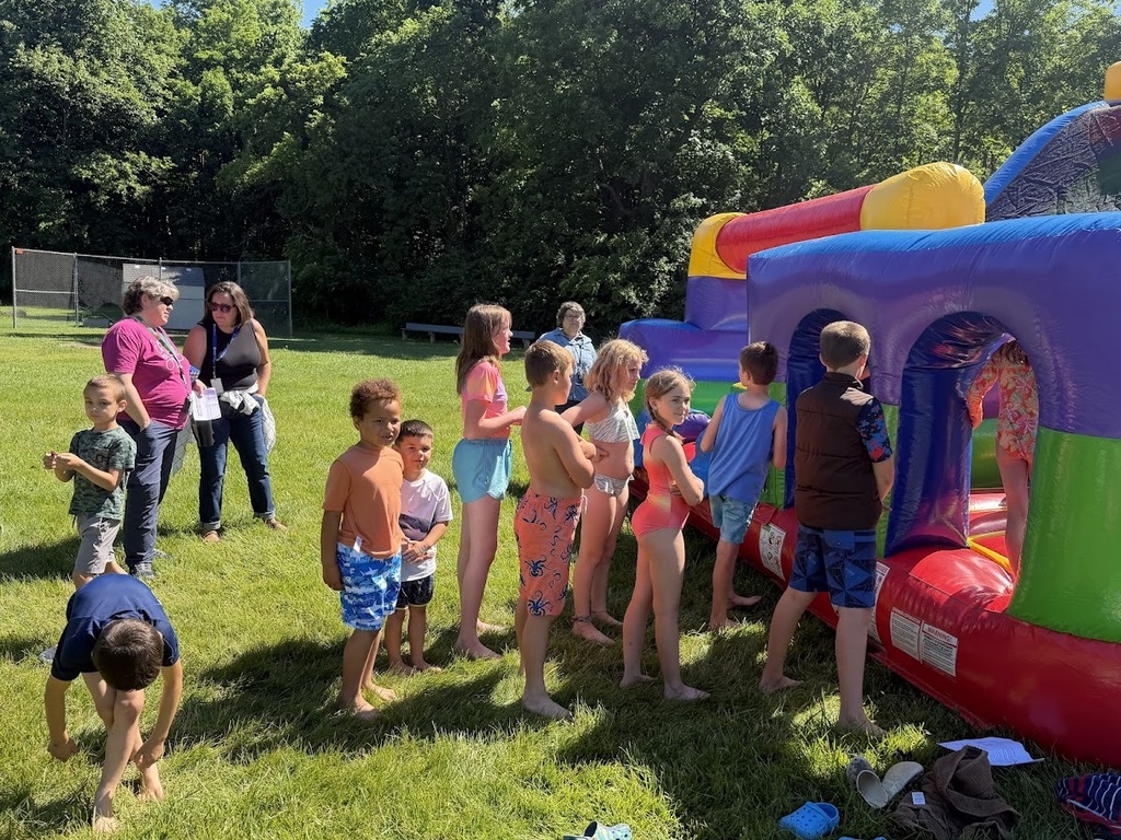 Students line up to take turns on a colorful inflatable obstacle course during a school field day while staff members supervise nearby.