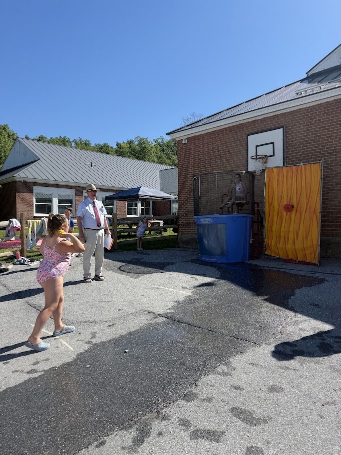A student takes aim at a dunk tank target while a staff member stands nearby supervising the activity on a sunny day outside the school.