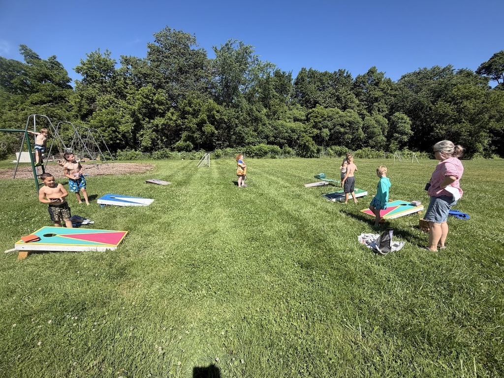 Students play cornhole on a grassy field near the playground while a staff member watches. Multiple cornhole boards are set up for friendly competition during field day.