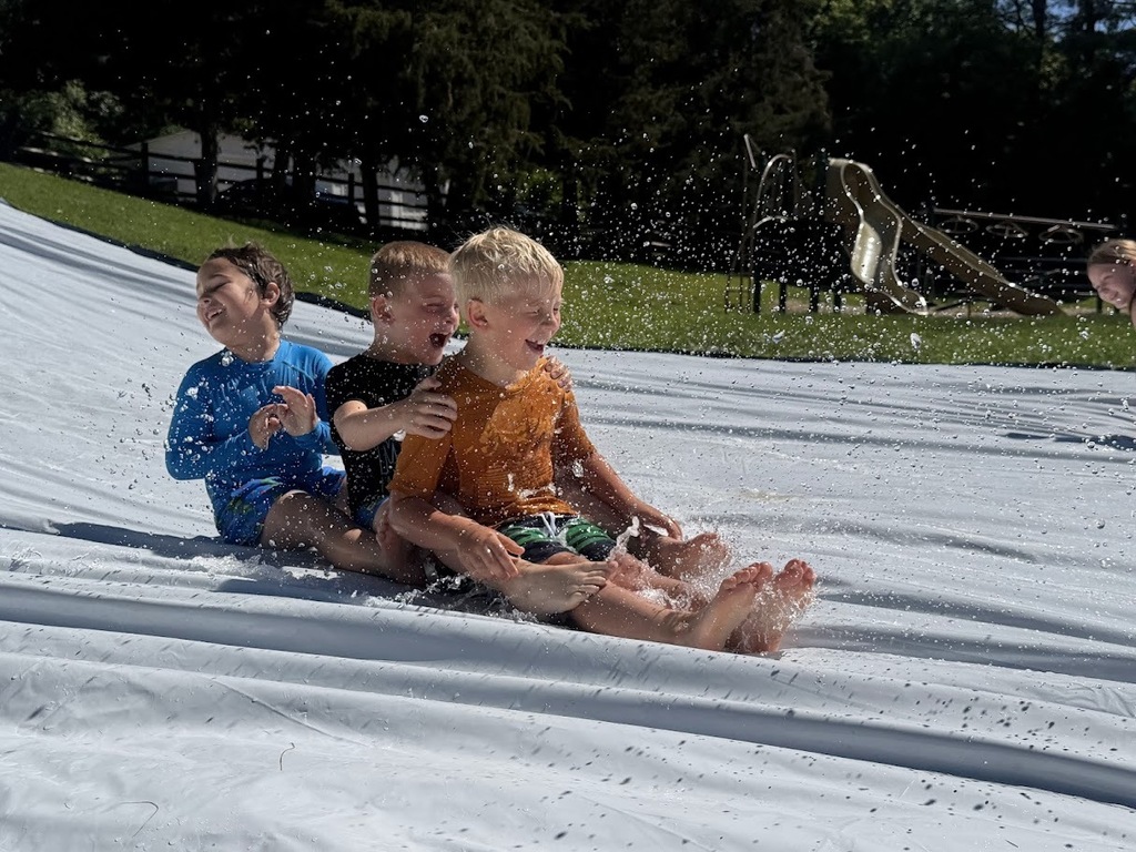 Three young students slide through splashing water on a slip-and-slide, laughing as they race down the hill during an outdoor field day.