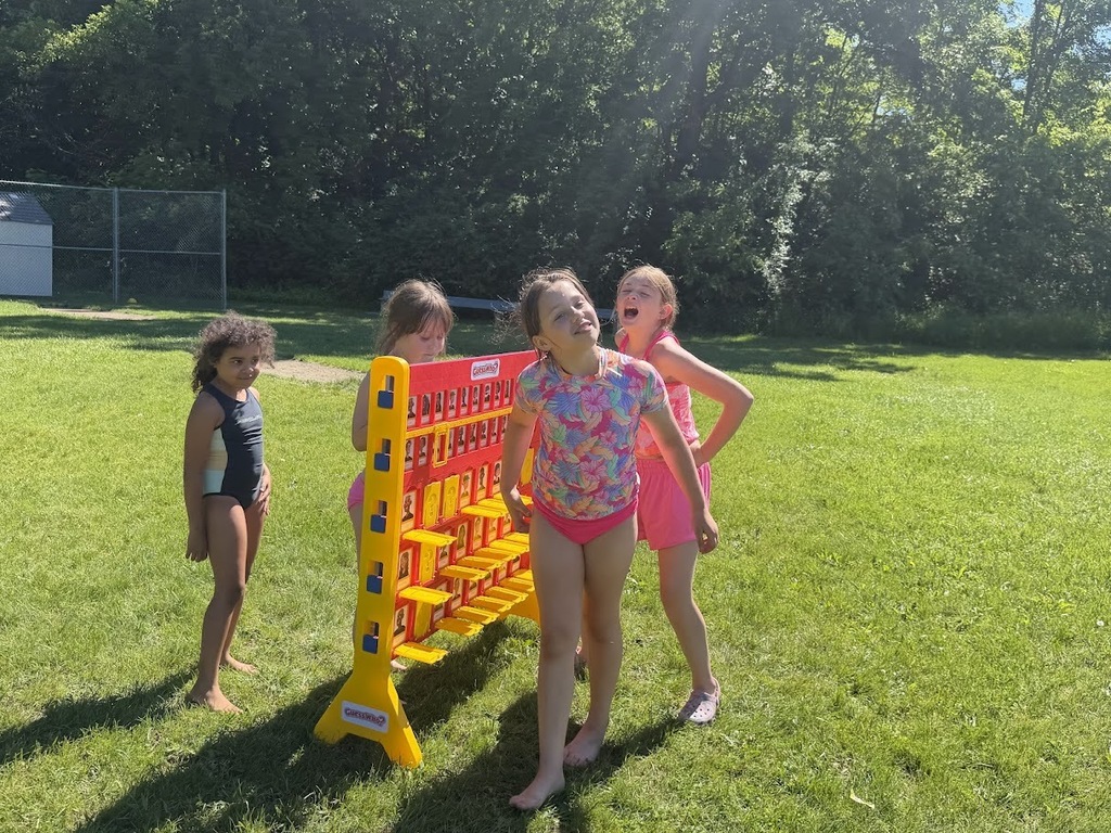 Students play a giant outdoor game of Guess Who? on the grass during a school field day. Several children stand around the oversized game board waiting for their turns.