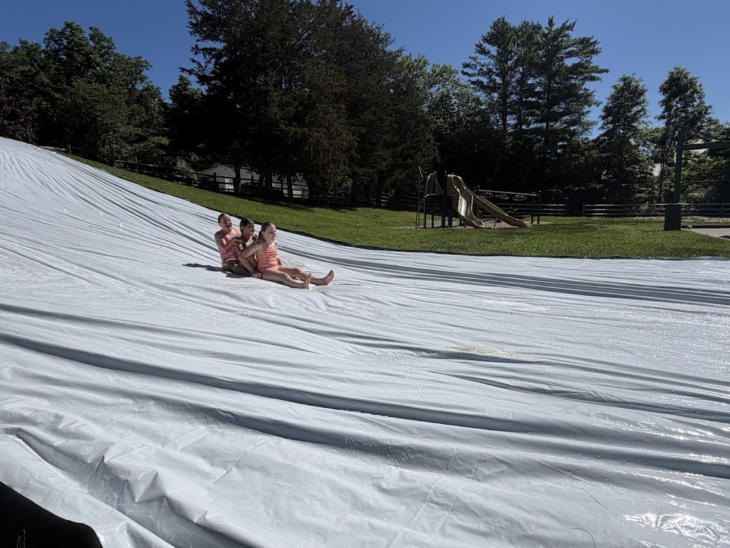 Three students ride together down a giant slip-and-slide, smiling and laughing as they glide across the wet surface.