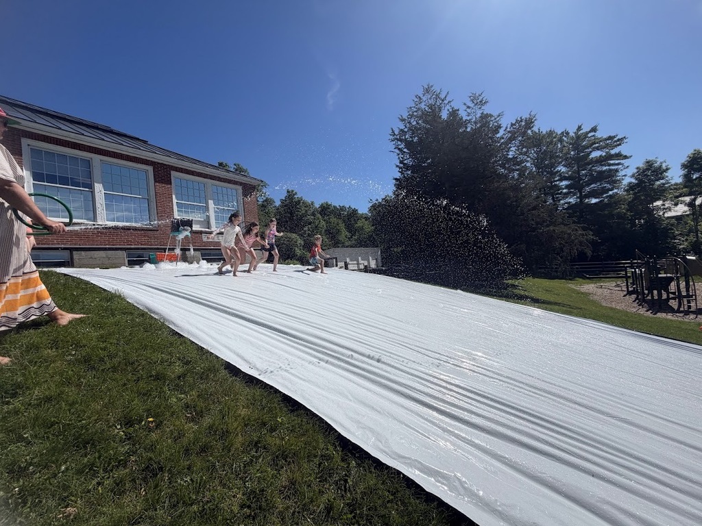 Students run and slide down a large homemade slip-and-slide spread across a grassy hill while water sprays overhead on a sunny day.