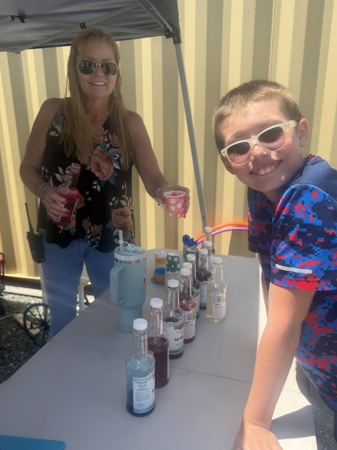 A student smiles at a flavored ice station while a staff member prepares a colorful frozen treat under a canopy tent.