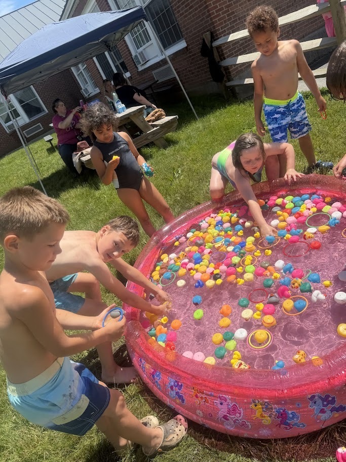 Students gather around a small inflatable pool filled with floating toys and rings, reaching into the water during a carnival-style field day game.